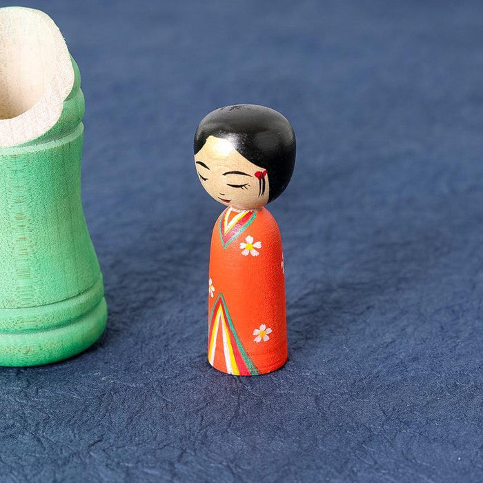 Kokeshi wooden doll figure with red kimono and floral design beside green bamboo case | Tsuchiyu, Fukushima