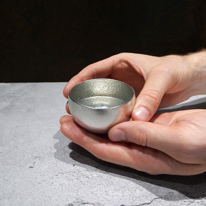Hands cradling a small silver tin sake cup with textured interior | Nousaku, Takaoka